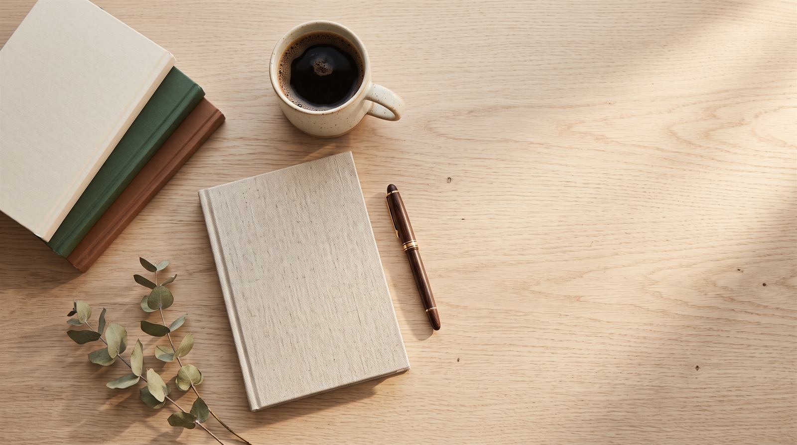 A writer's desk seen from above: a closed linen notebook, a fountain pen, a mug of black coffee, a stack of paperbacks and a sprig of dried eucalyptus on pale oak, lit by warm morning light.
